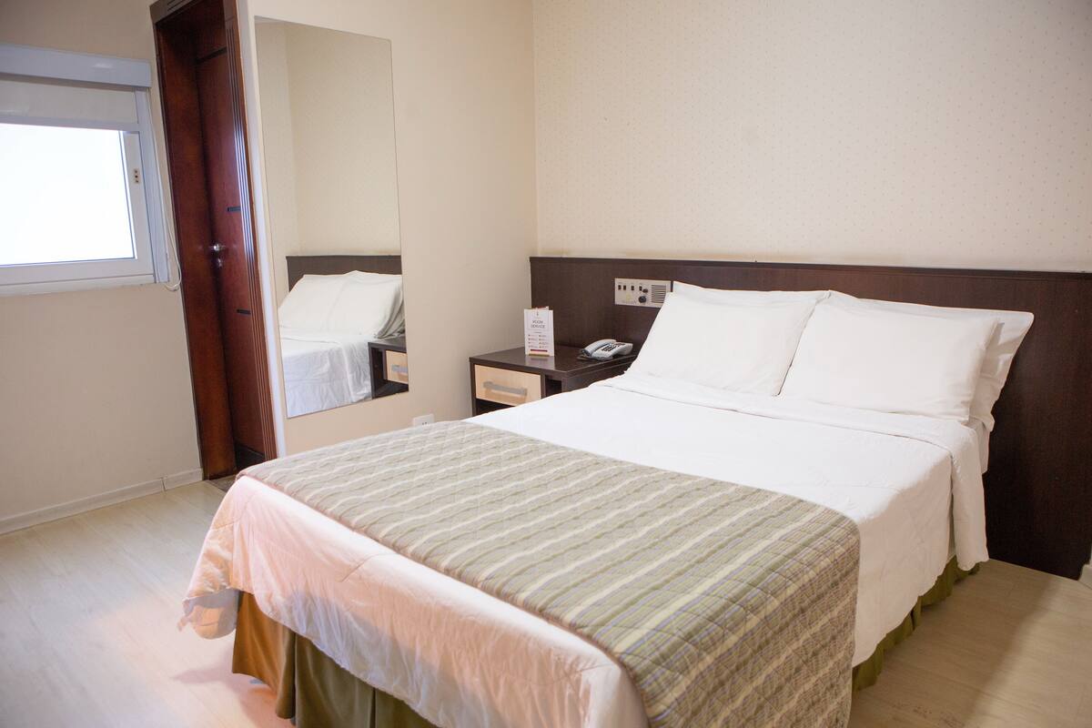 Una habitación de hotel con cama, mesita de noche, espejo y ventana.