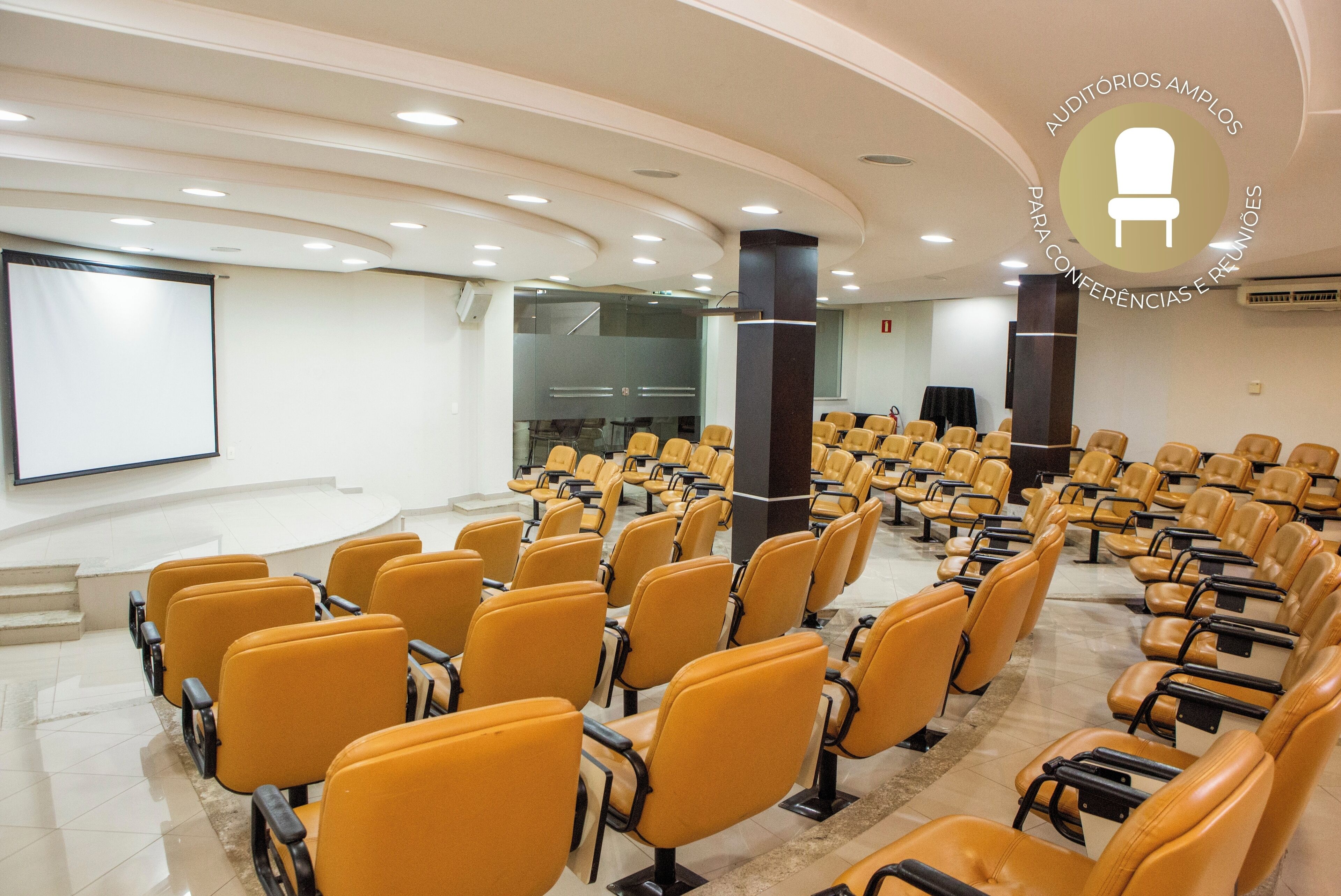 A conference room with rows of yellow chairs, a large screen, and a podium.