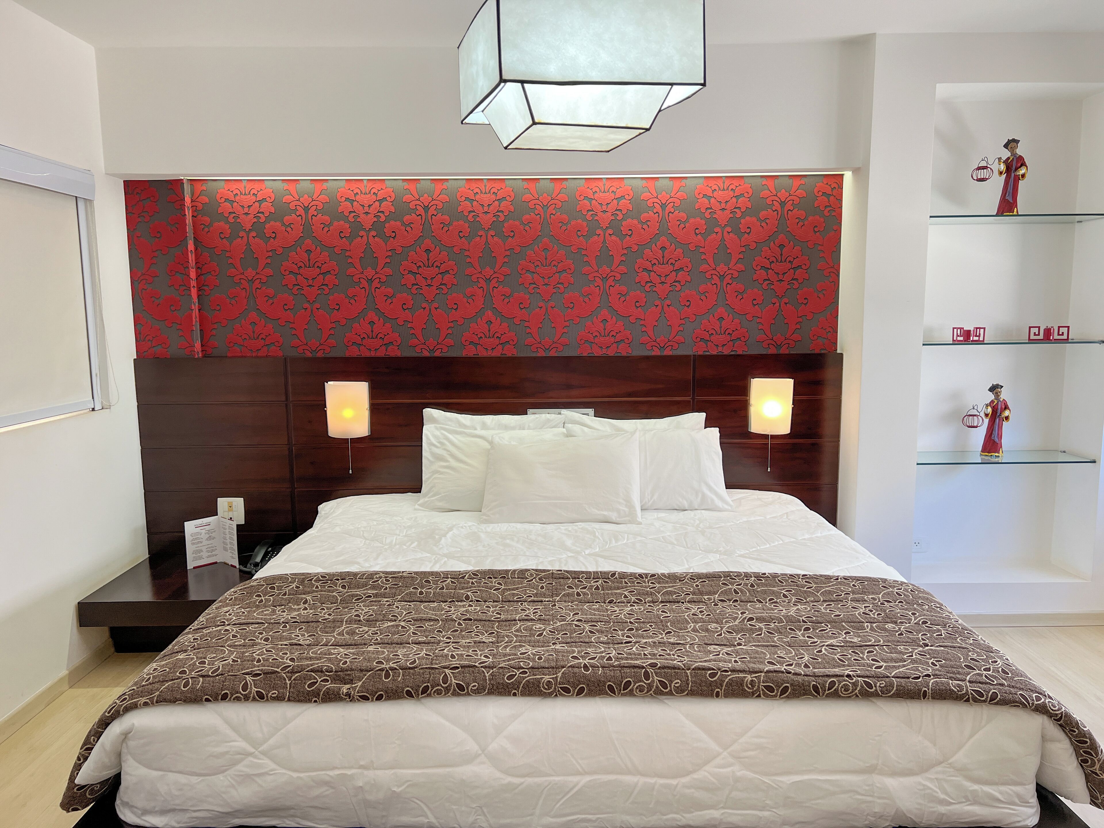 Una cama bien hecha con ropa de cama blanca y un plaid marrón estampado, un cabecero de madera y papel tapiz rojo y gris, además de lámparas de noche.