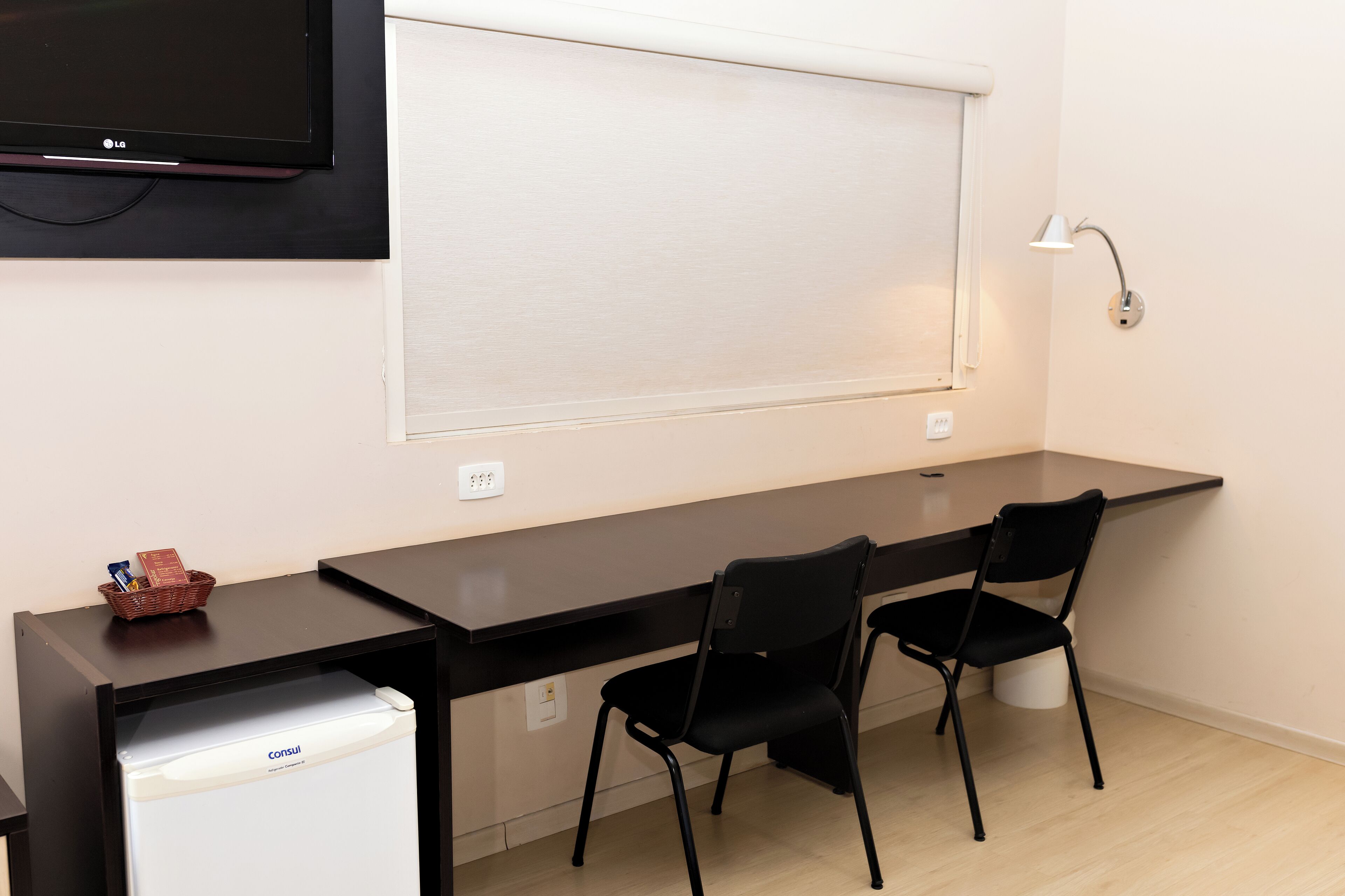 A small office space with a desk, two chairs, a flat-screen TV, a white mini-fridge, a lamp, and a window with blinds.