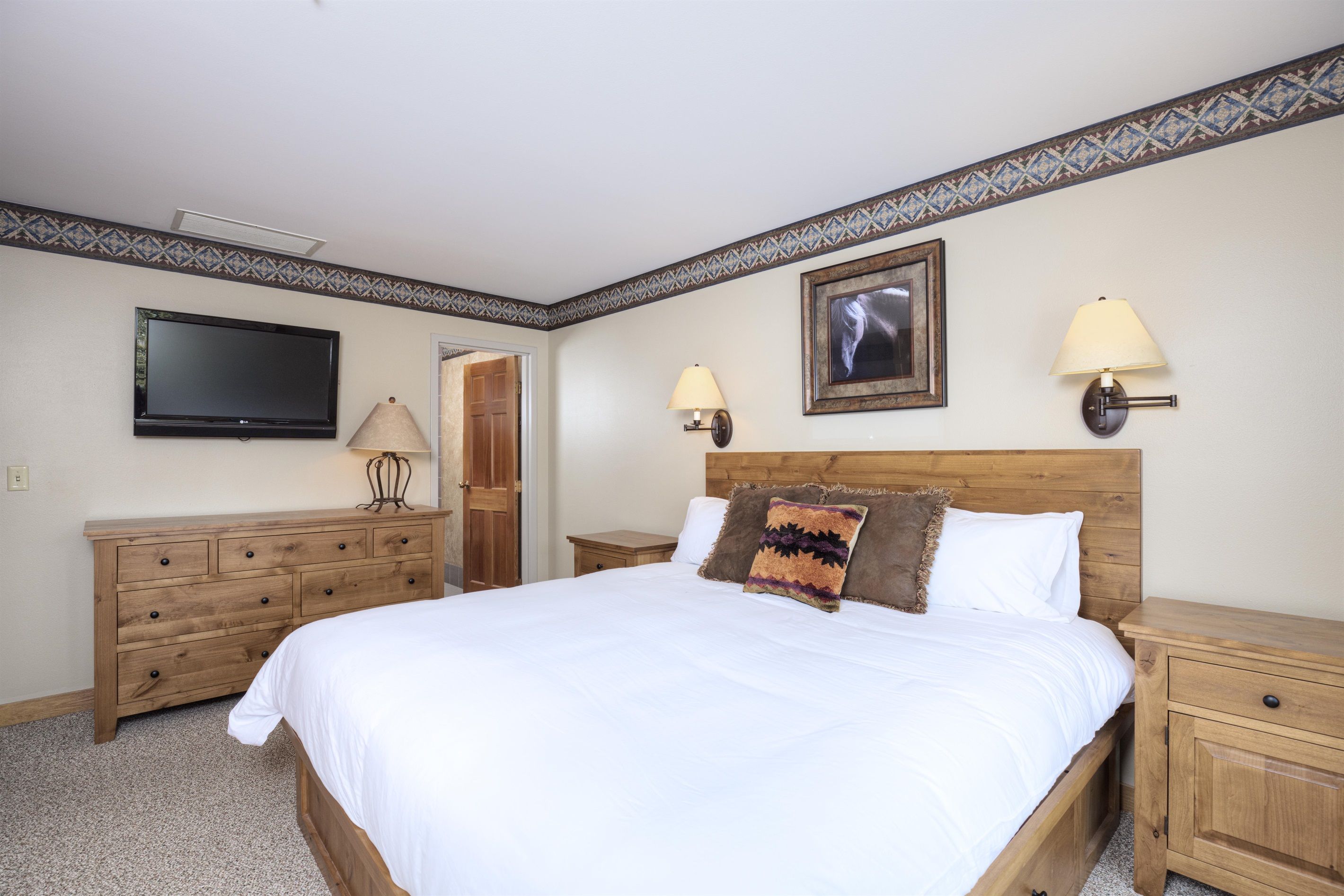 Un dormitorio con una cama grande, mesitas de noche de madera, un aparador, un televisor de pantalla plana, dos lámparas de pared y un cuadro en la pared.