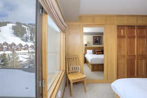 A hotel room with a large window offering a view of a snowy mountain and cabins. The room features wooden furniture, a bed, a chair, and a wardrobe.