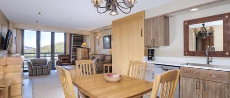 A modern kitchen with wooden cabinets, a dining table with chairs, and a large window offering a view of the outdoors.