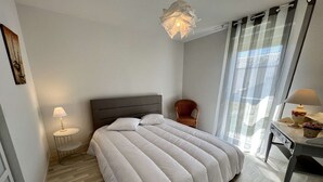A modern bedroom with a large bed, a bedside table with a lamp, a chair, and a view of the outdoors through sheer curtains. - Etel - 2-room apartment - 43 sqm - Wifi - Classified 3* (ETEL)