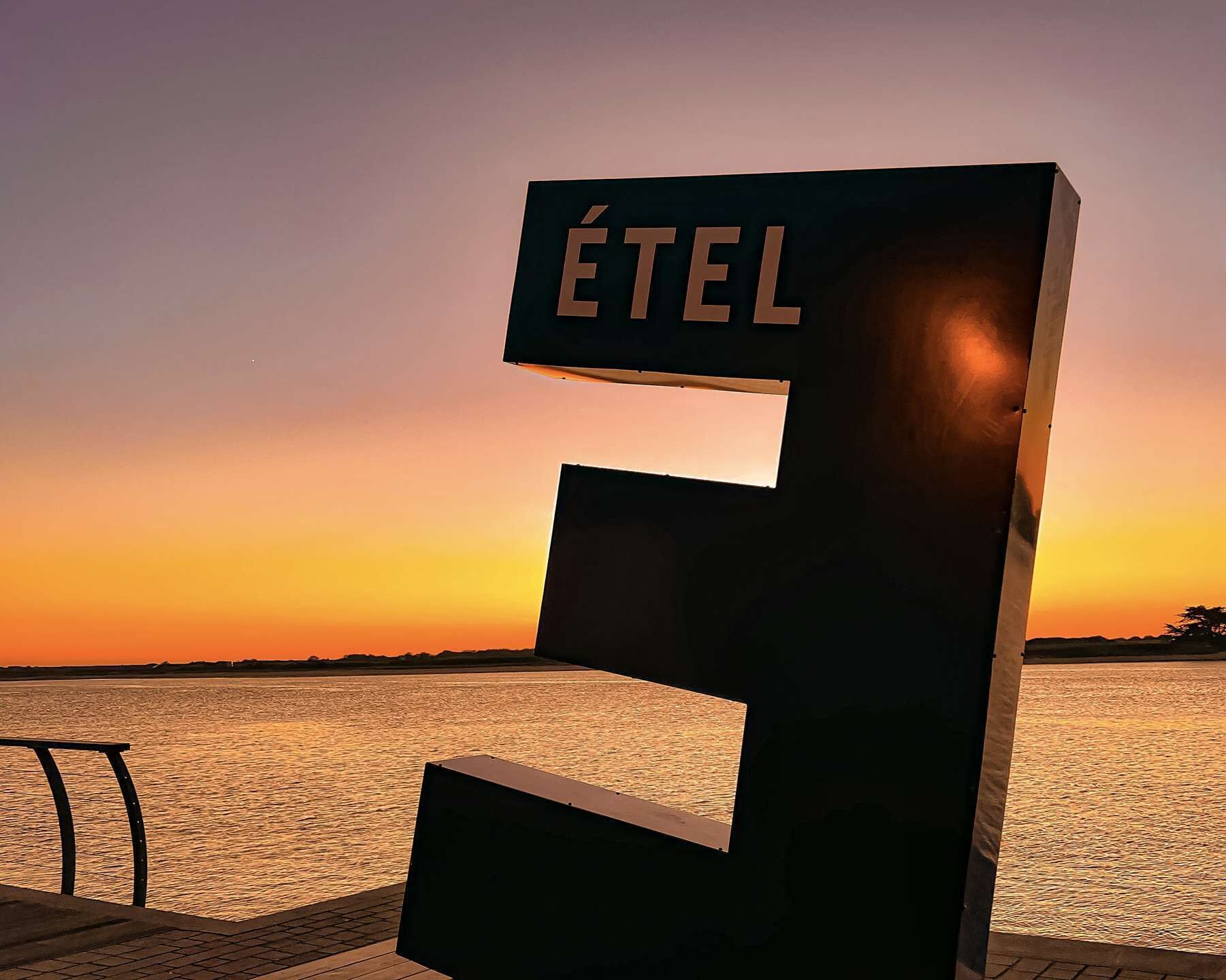 A large, bold, three-dimensional letter 'E' with the word 'ÉTEL' on it, set against a sunset backdrop over a body of water.