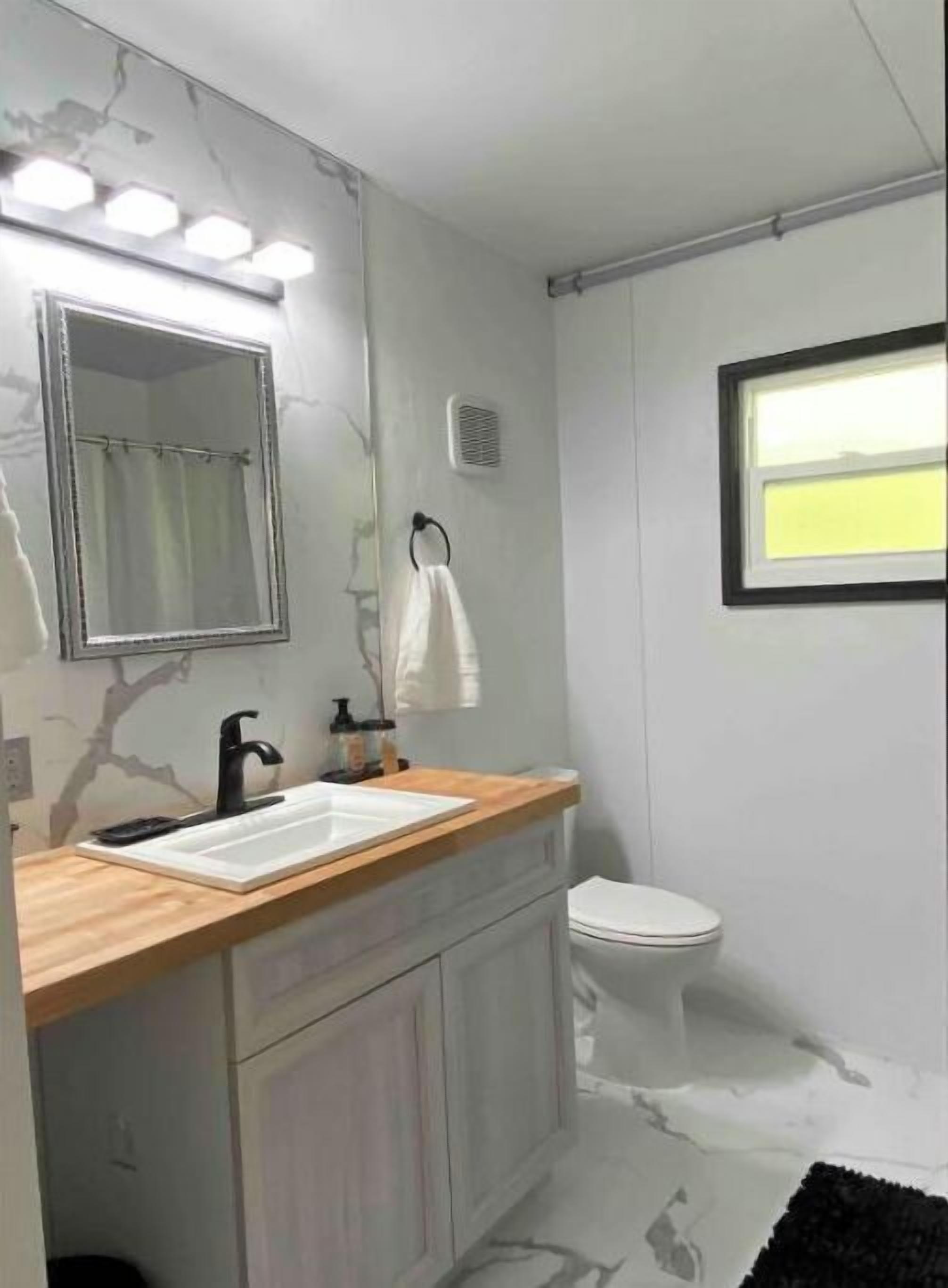 A bathroom with a wooden vanity, a white sink, a black faucet, a mirror, a towel ring, a toilet, and a window with a view of the outside.