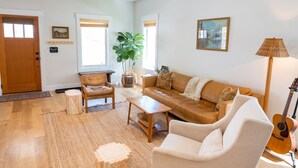 Un salón amplio con piso de madera, un sofá de cuero marrón, un sillón blanco, una mesa de centro y una guitarra.