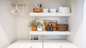Un baño con muebles blancos, un perchero, una repisa con artículos de aseo y una planta en maceta.