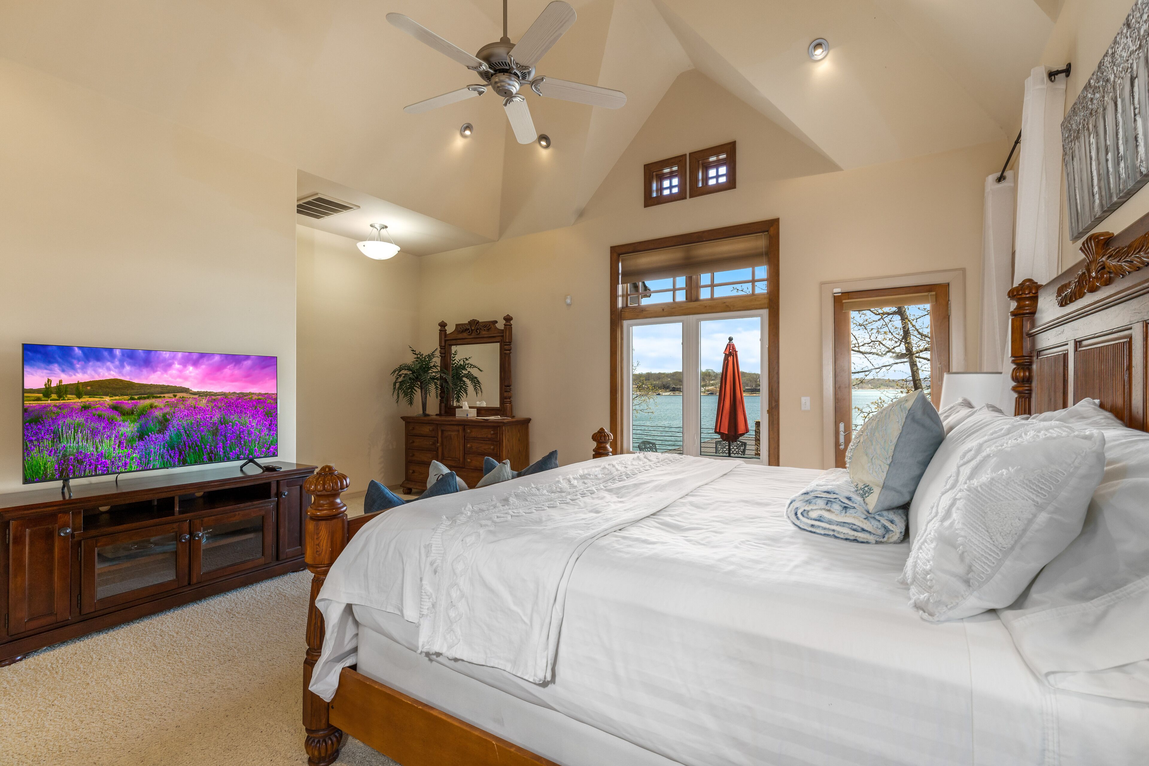 A bedroom with a large bed, a ceiling fan, a TV, a dresser, and a view of the outdoors.