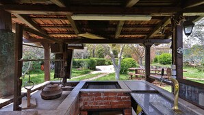Uma área de bar ao ar livre com churrasqueira de tijolos, vigas de madeira e vista para um jardim.