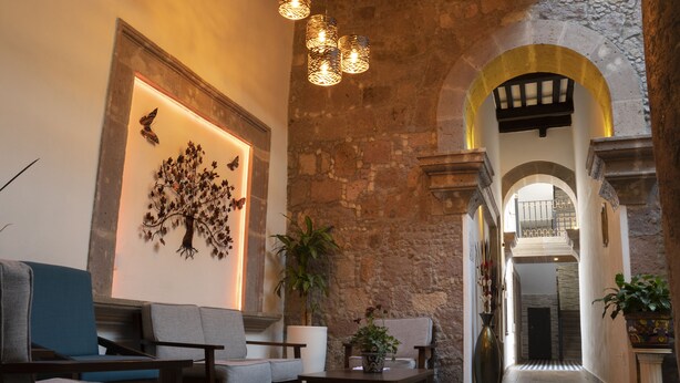 A lobby with a stone wall, a blue sofa, a wooden coffee table, and a potted plant. - Hotel Don Carlos (Morelia)