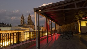 An outdoor patio with a covered seating area, a stone wall, and a view of two historic towers in the background. - Hotel Don Carlos (Morelia)