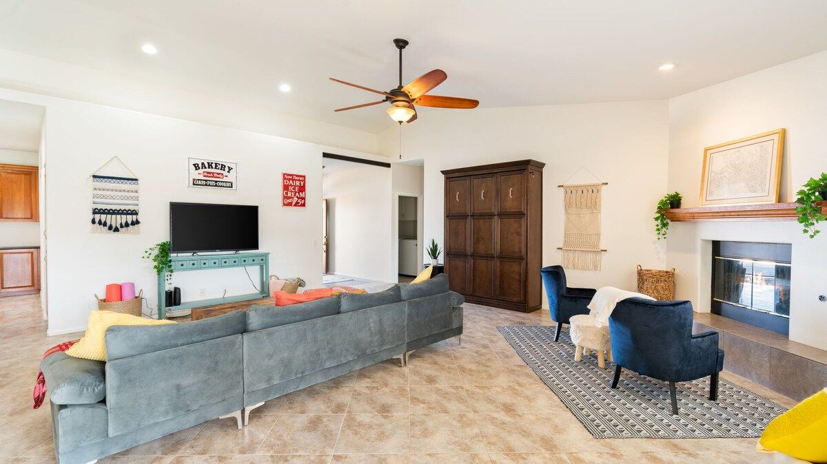 A living room with a sofa, armchairs, a television, a ceiling fan, a fireplace, and a kitchen area in the background.