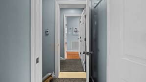 A hallway with a door leading to a room with a window, a door with a handle, and a wall-mounted light switch. - Shore Thing West -Spacious Beachfront Oasis & Pool - 5BR (Emerald Isle)
