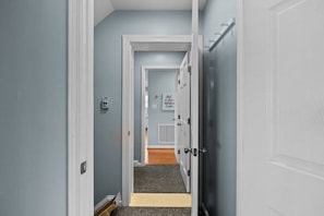 A hallway with a door leading to a room with a window, a door with a handle, and a wall-mounted light switch. - Shore Thing West -Spacious Beachfront Oasis & Pool - 5BR (Emerald Isle)