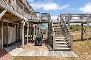 A wooden deck with a staircase leading to a covered area, surrounded by a natural landscape. - Shore Thing West -Spacious Beachfront Oasis & Pool - 5BR (Emerald Isle)