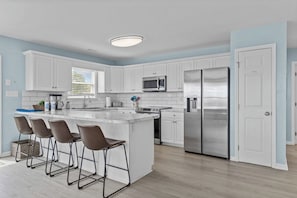 A modern kitchen with white cabinetry, stainless steel appliances, and a central island with bar stools. - Shore Thing West -Spacious Beachfront Oasis & Pool - 5BR (Emerald Isle)