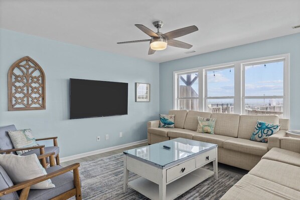 A living room with a ceiling fan, a flat-screen TV, a beige sofa, and a glass coffee table. - Shore Thing West -Spacious Beachfront Oasis & Pool - 5BR (Emerald Isle)
