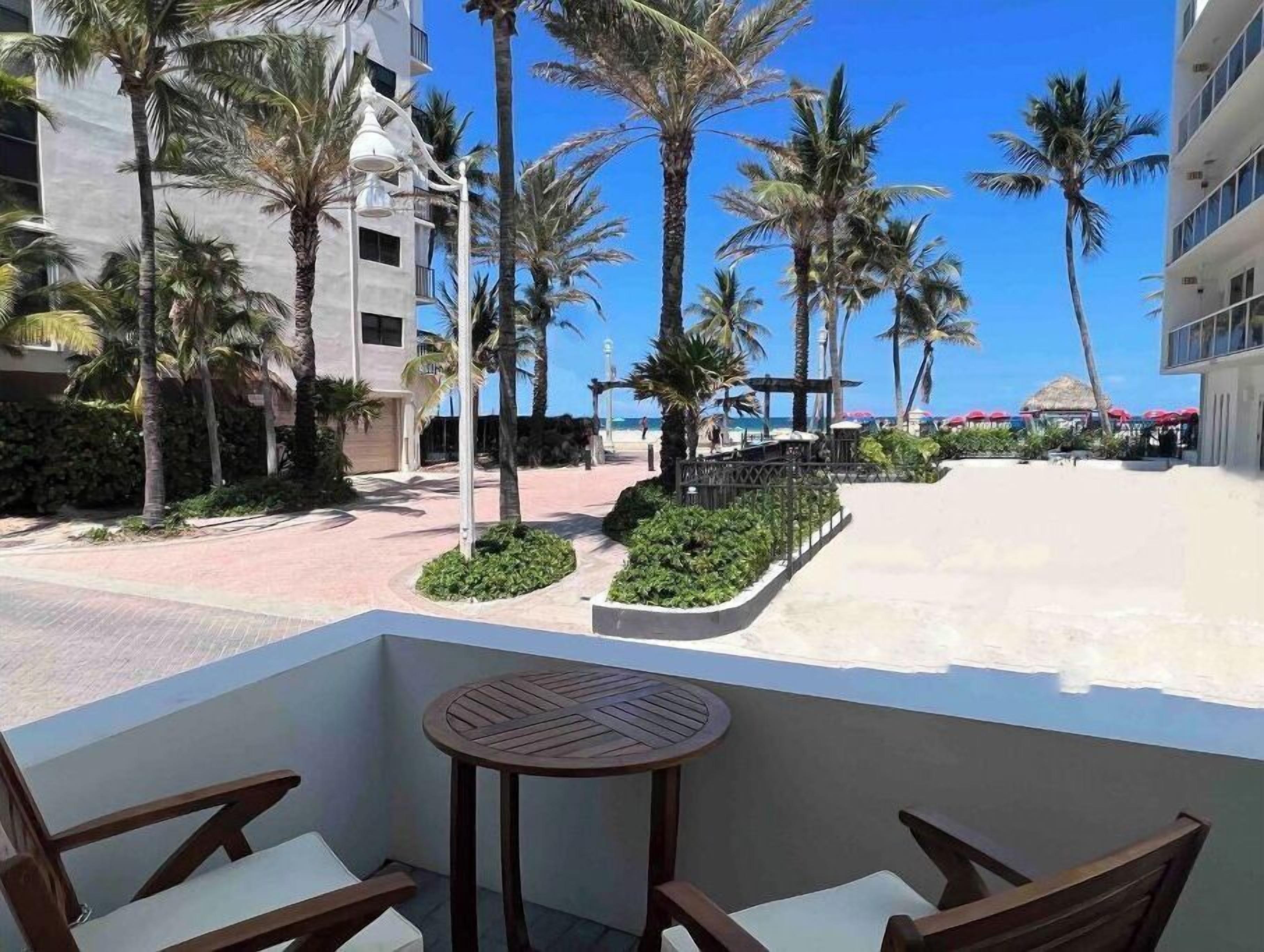 Un balcón con una mesa redonda y dos sillas, con vista a una playa con palmeras y un edificio al fondo.