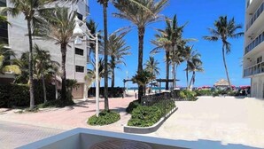 Un balcón con una mesa redonda y dos sillas, con vista a una playa con palmeras y un edificio al fondo.