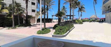 A balcony with a round table and two chairs, overlooking a beach with palm trees and a building in the background.