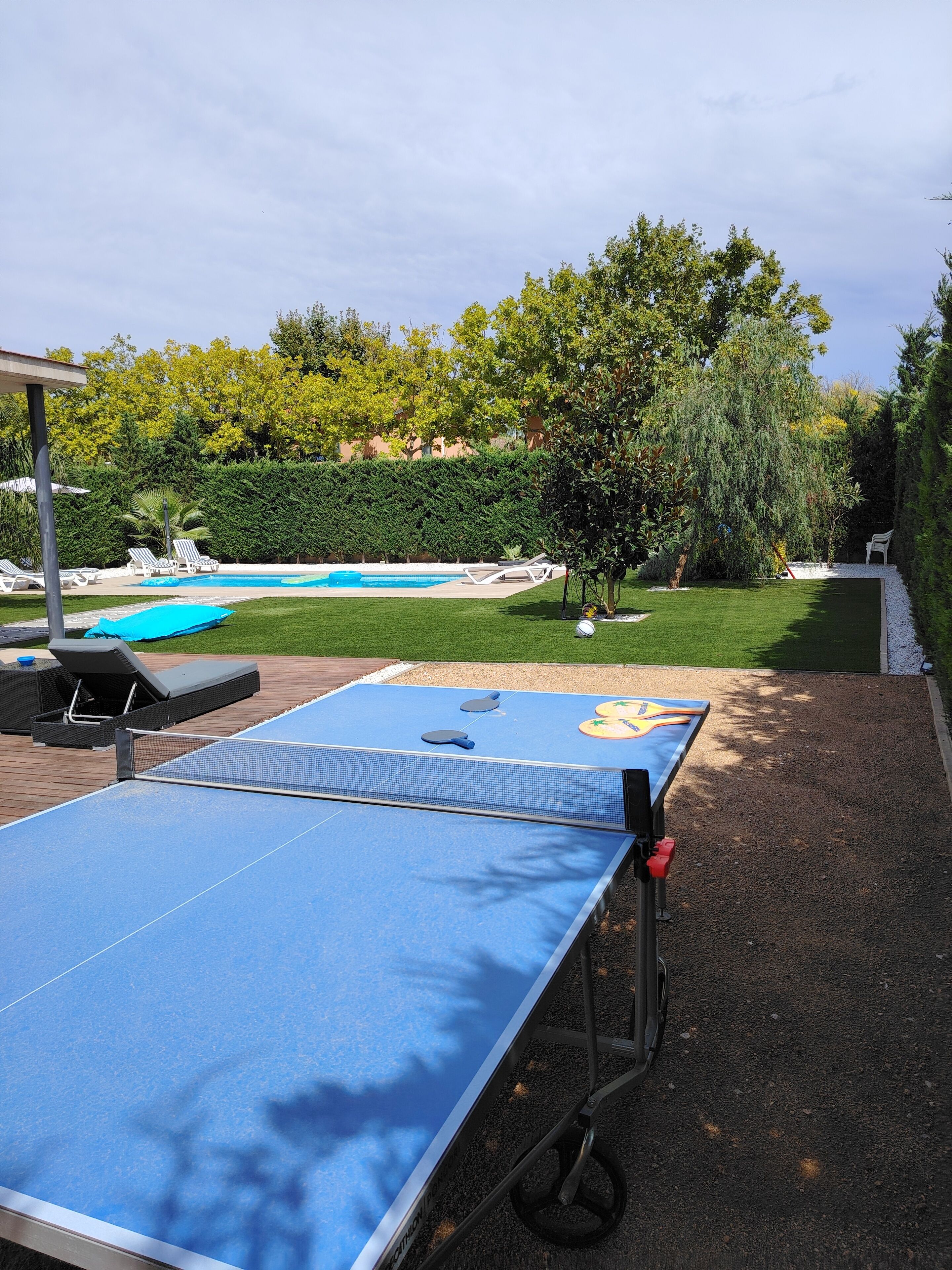 An outdoor ping pong table with a net, surrounded by a pool and greenery.
