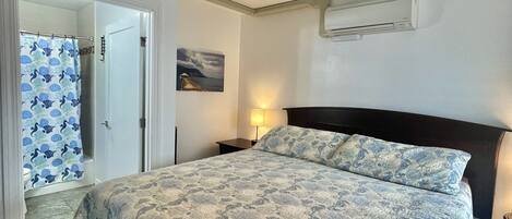 Un dormitorio con cama, ventilador de techo, equipo de aire acondicionado, mesita de noche y baño con cortina de ducha.