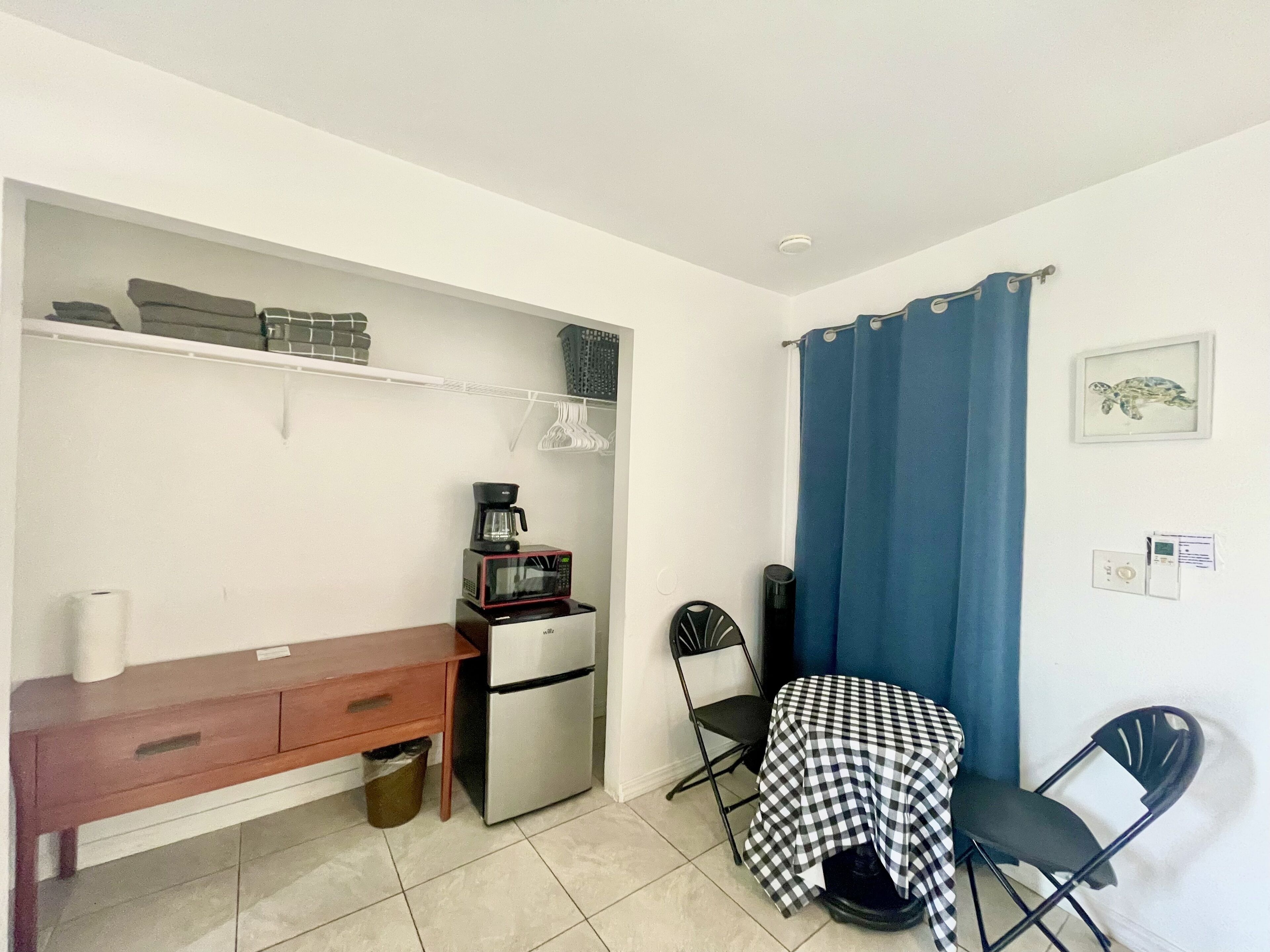 A compact kitchenette with a refrigerator, microwave, and coffee maker. A small table with a checkered tablecloth and two chairs are positioned near a blue curtain.