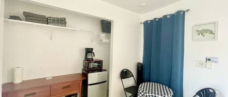 A compact kitchenette with a refrigerator, microwave, and coffee maker. A small table with a checkered tablecloth and two chairs are positioned near a blue curtain.