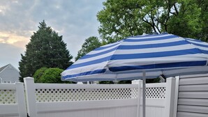 A patio with a round glass table and four chairs, a blue and white striped umbrella, and a white picket fence. - The Heart of Raleigh (Raleigh)