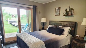 A bedroom with a large bed, a wooden headboard, two bedside lamps, a small table, and a view of a garden through a sliding door. - Modern NEWLY renovated 1 bedrm, WIFI, Pool, new kitchen Summer of 24!  sleeps 4. (Mancelona)