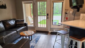 A living room with a leather couch, a coffee table, and a TV mounted on the wall. There are two doors leading to an outdoor area with a lawn and a chair. - Modern NEWLY renovated 1 bedrm, WIFI, Pool, new kitchen Summer of 24!  sleeps 4. (Mancelona)