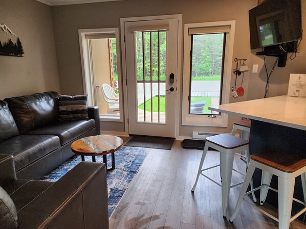 A living room with a leather couch, a coffee table, and a TV mounted on the wall. There are two doors leading to an outdoor area with a lawn and a chair.