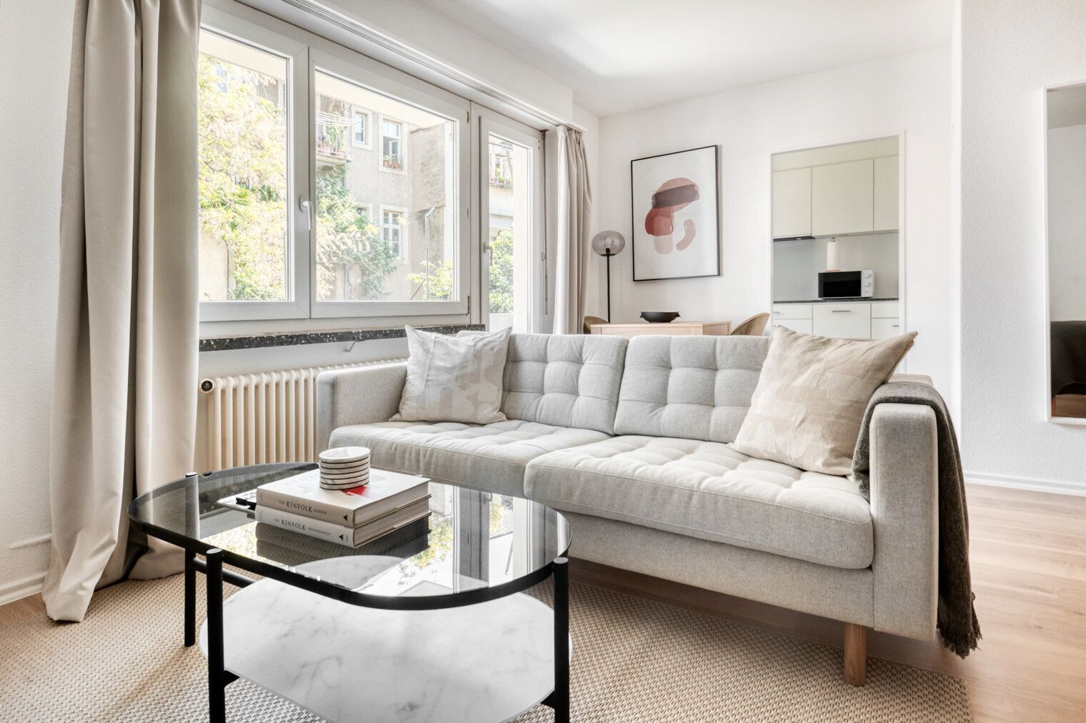 A modern living room with a grey sofa, a glass coffee table with books, and a view of the outside through large windows.