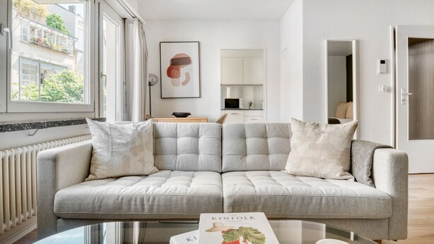 A modern living room with a grey sofa, a glass coffee table, and a book titled "The Infolk Garden". - Premium Iselin 1 Room w/ Storage, 1min to Local Market, by Blueground (Basel)