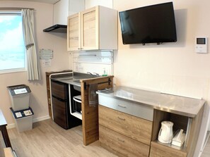 A compact kitchen with wooden cabinets, a stainless steel sink, a built-in oven, and a flat-screen TV mounted on the wall.
