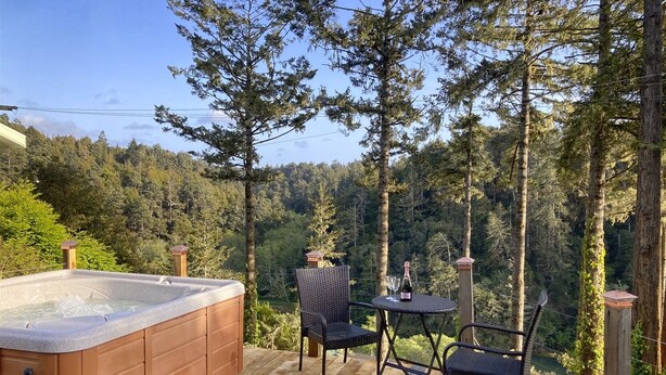 Un jacuzzi en una terraza de madera con dos sillones de mimbre y una mesa, rodeado de árboles altos y una ladera boscosa.