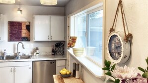 Una cocina con armarios blancos, lavavajillas de acero inoxidable, tabla de madera con limones y ventana con vistas al exterior.