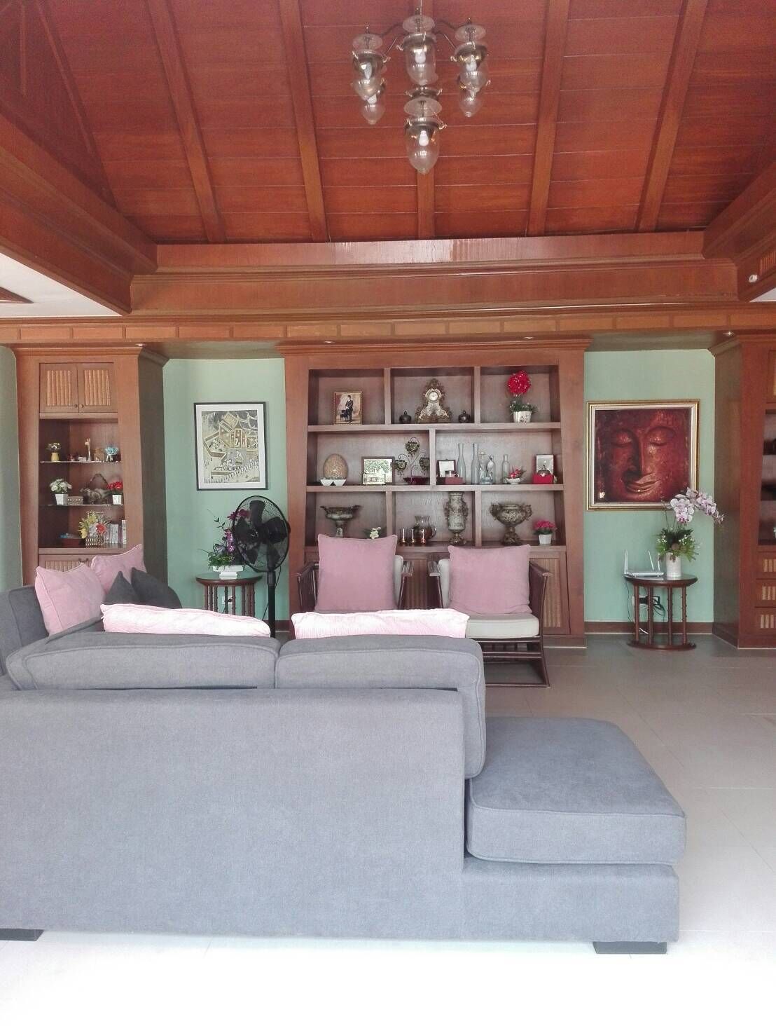 A spacious living room with a large grey sectional sofa, wooden ceiling, and a wall-mounted shelf with decorative items.