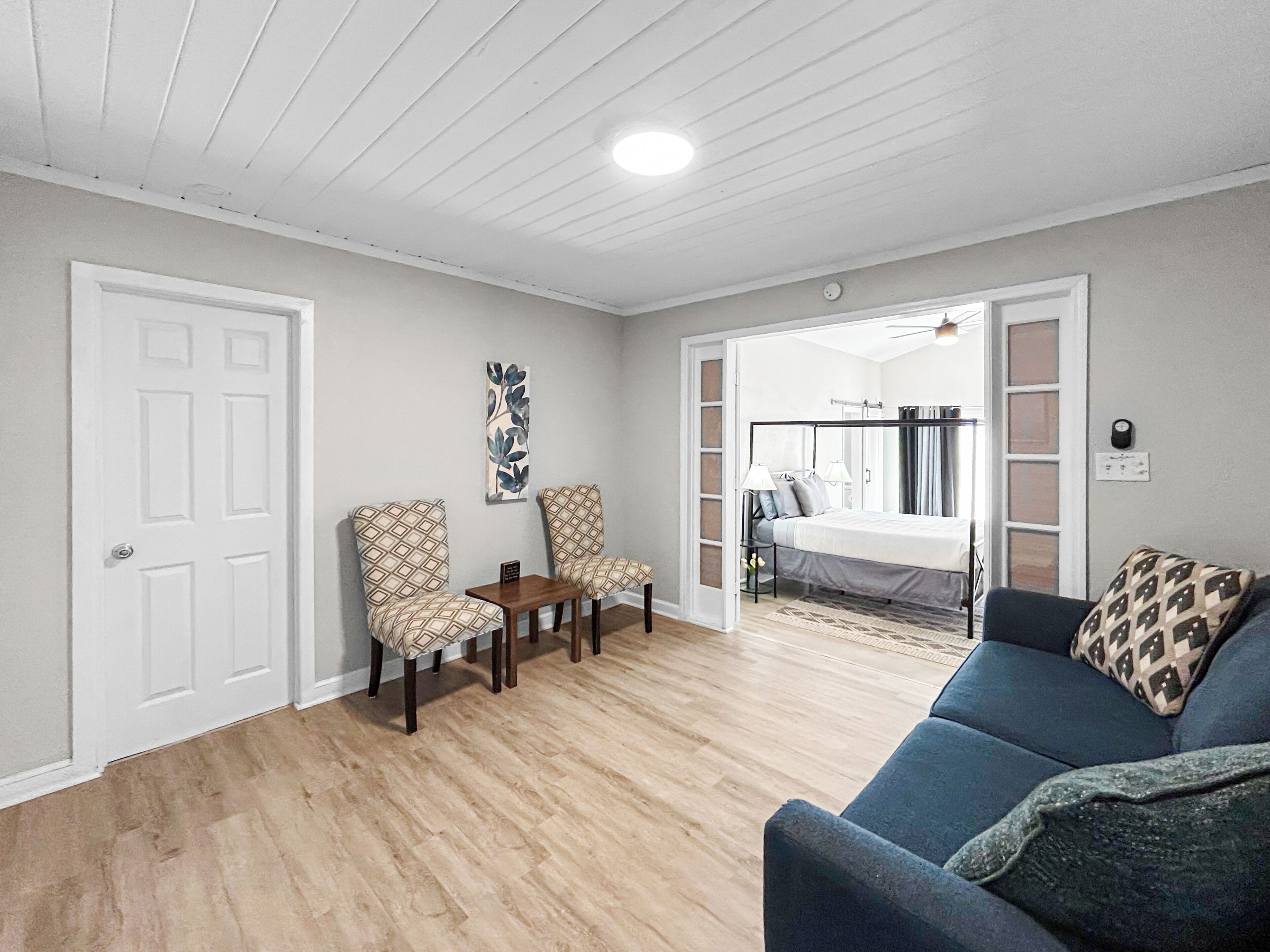 A room with a bed, a blue sofa, two patterned chairs, a small table, and a door.