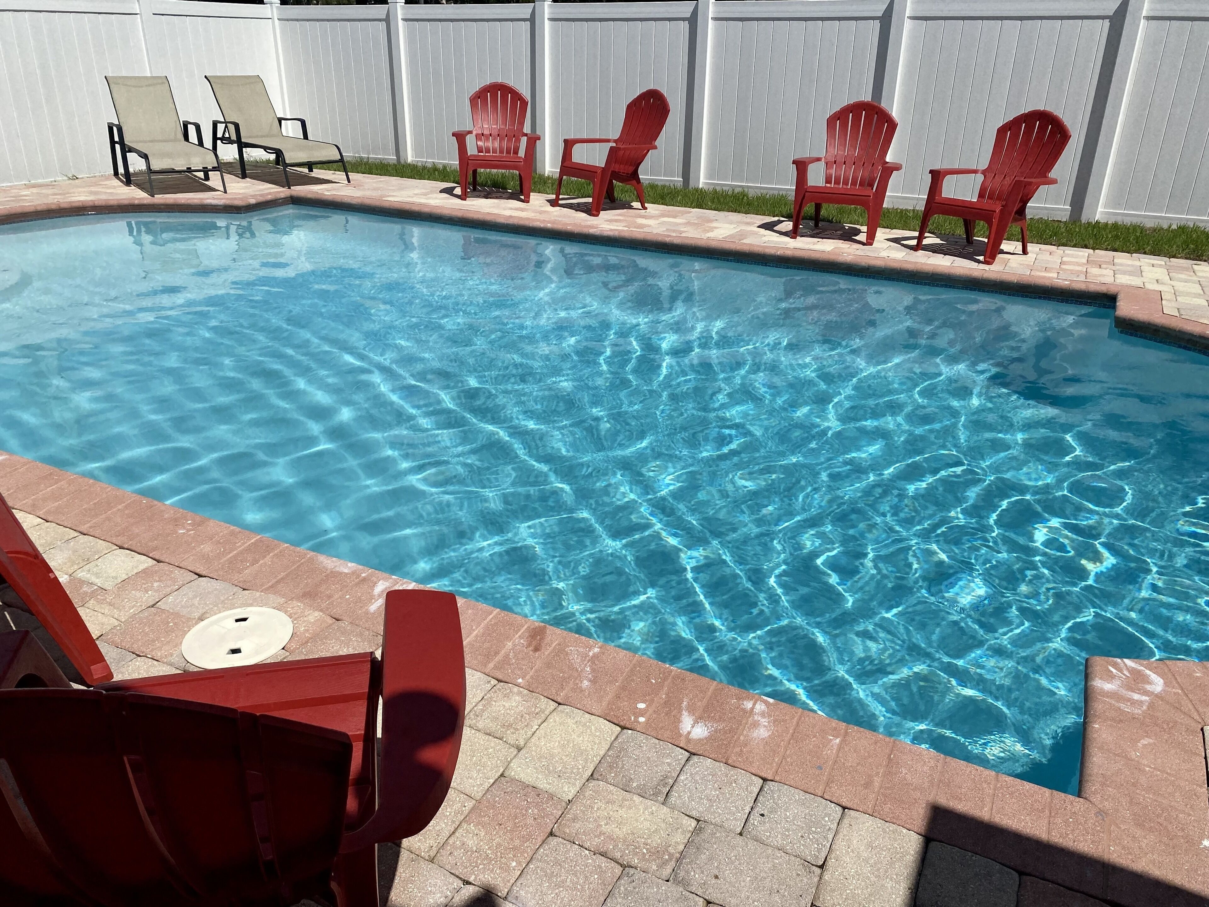 Une piscine à l’eau d’un bleu cristallin, entourée d’une terrasse en briques et de chaises Adirondack rouges.