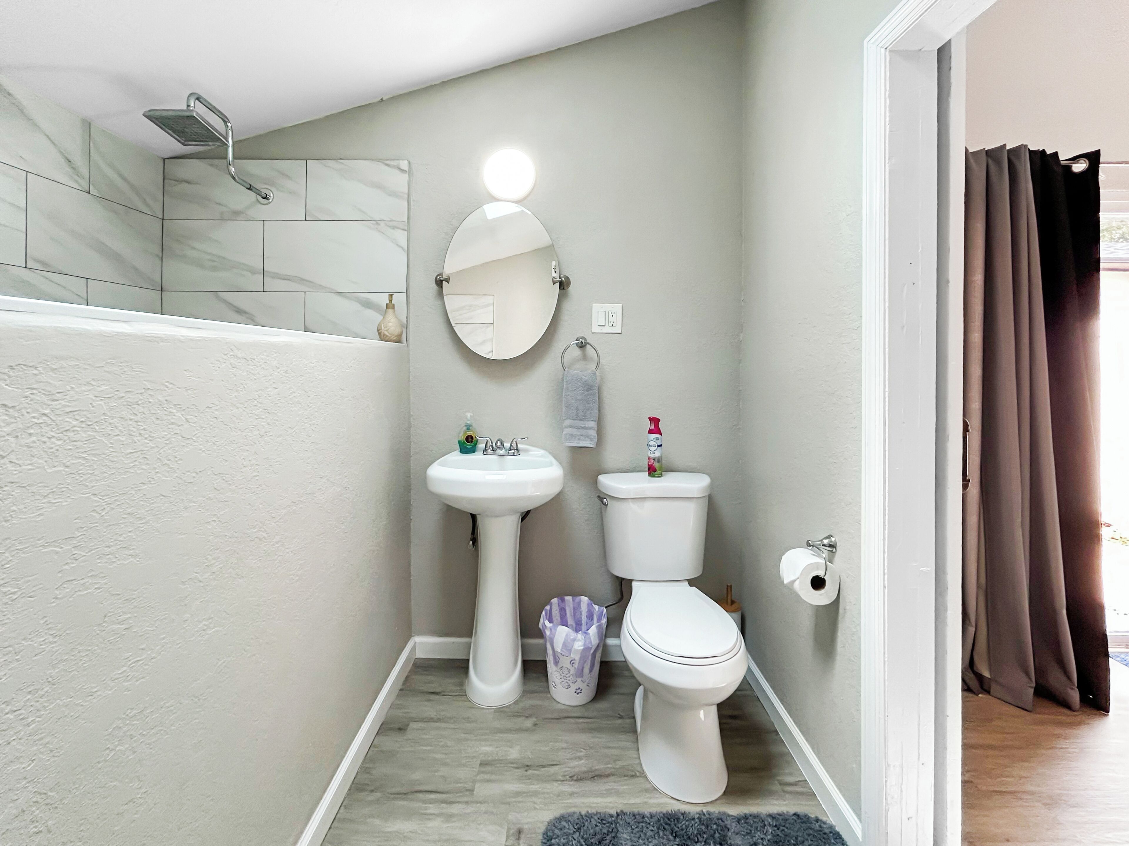 A bathroom with a white sink, toilet, and shower. There is a mirror with a light above it, and a small trash bin is visible.