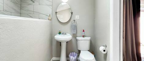 Et badeværelse med en hvid vask, toilet og brusebad. Der er et spejl med lys over, og en lille skraldespand er synlig.