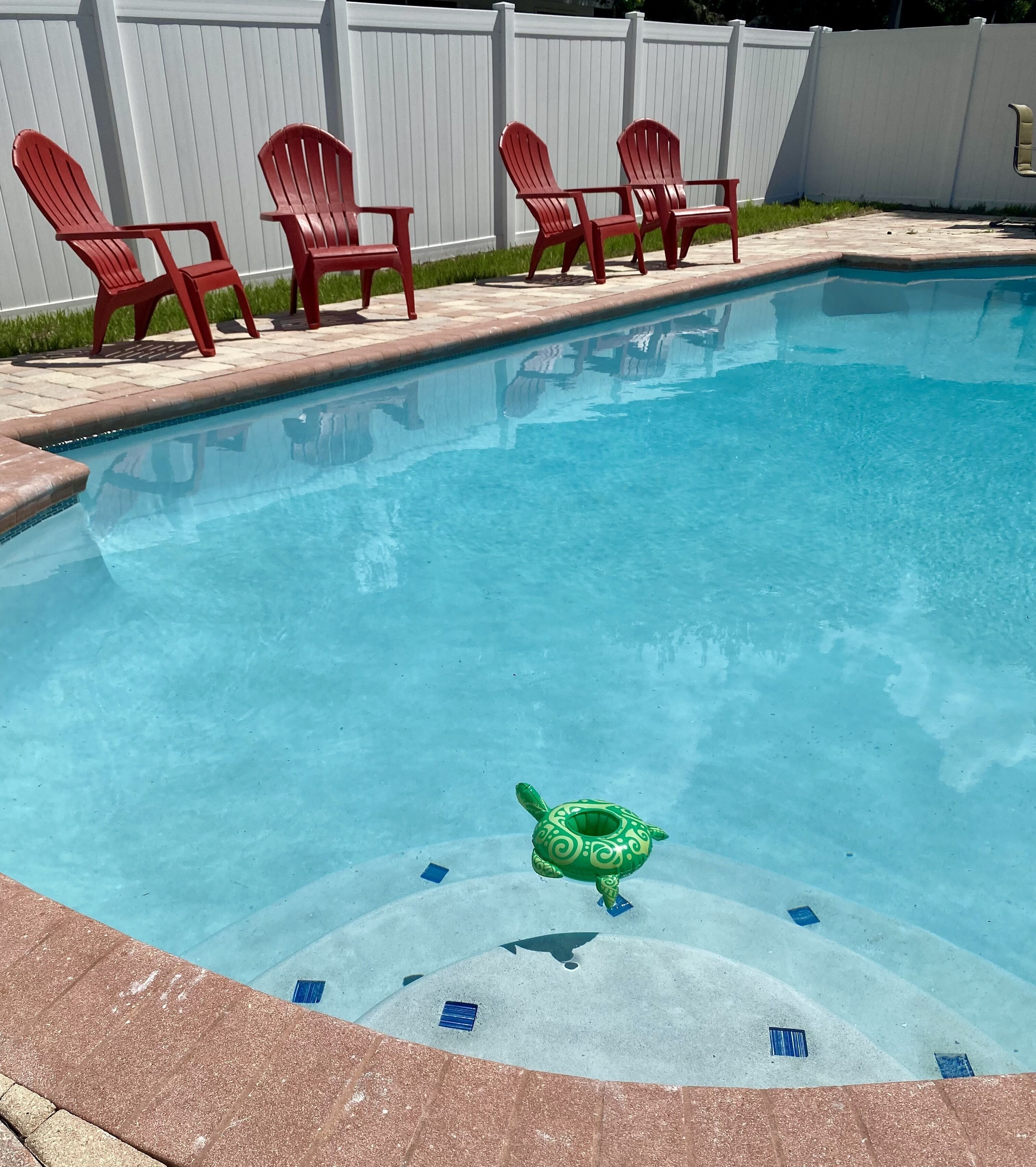 A swimming pool with a green inflatable toy, red chairs, and a white fence in the background.