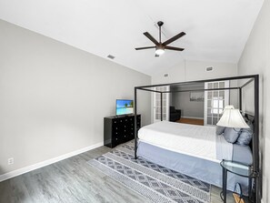 A bedroom with a four-poster bed, a ceiling fan, a TV on a stand, and a bedside table with a lamp.
