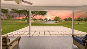 En overdækket terrasse med ventilator, udsigt til en park med træer og en solnedgang.