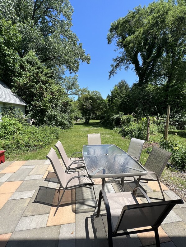 A patio with a glass-top table and metal chairs, surrounded by a stone-tiled floor and lush greenery. - Modern Farmhouse, Sleeps 8, North of St. Joseph (Benton Harbor)