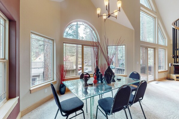Enjoy meals in this dining area with a glass-top table and four chairs, surrounded by large windows offering views of the surrounding trees and a distant building.