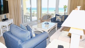 Ein Wohnzimmer mit einer Couch, einem Sessel und einem Couchtisch, großen Fenstern mit Meerblick und einem Flachbildfernseher auf einem Standfuß.