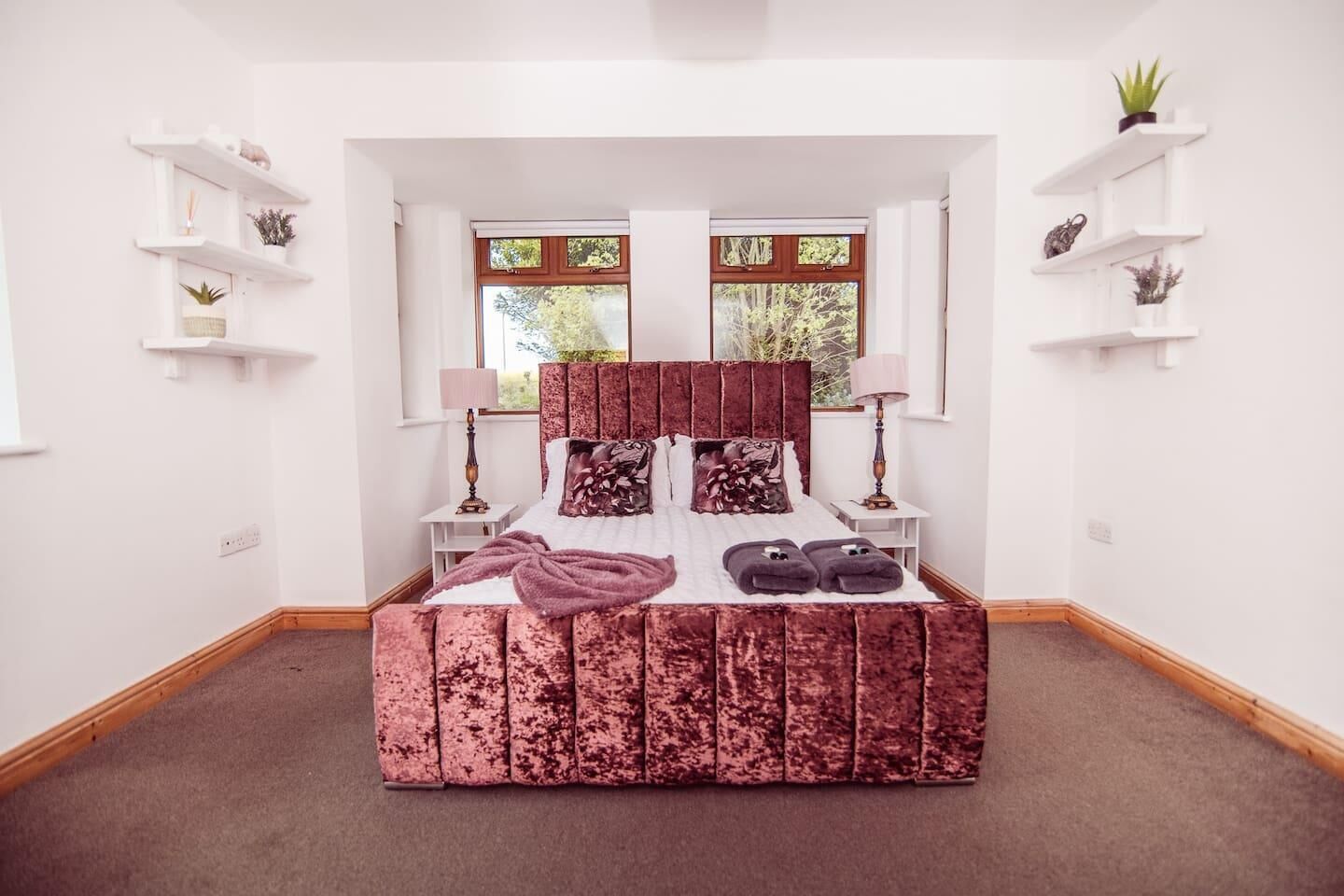 A bedroom with a large bed, two bedside tables, a window with curtains, and white shelving units with decorative items.